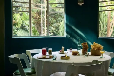 Round dining table set with food and drinks in a room with deep teal walls and large windows showing lush greenery outside