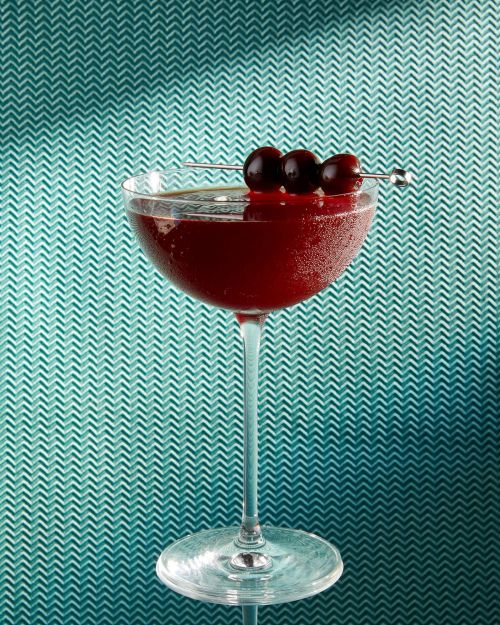 A stemmed cocktail glass filled with a dark red drink is garnished with three cherries on a metal pick, set against a teal chevron background.