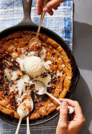 Overhead shot of a cast iron skillet containing a large chocolate chip cookie topped with a scoop of vanilla ice cream and several spoons.