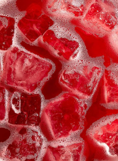 A close-up view shows ice cubes and bubbles in a vibrant red liquid, possibly a refreshing beverage, suggesting refreshment and coolness.