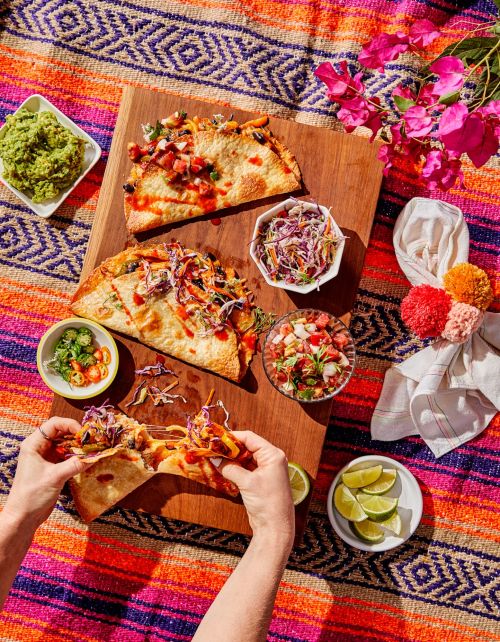 Overhead shot of someone holding a taco over a wooden board with various toppings and sides, set on a colorful striped blanket.