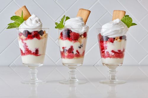 Three glass dessert cups filled with layers of berries, cream, and cookies, topped with mint, against a white brick background.