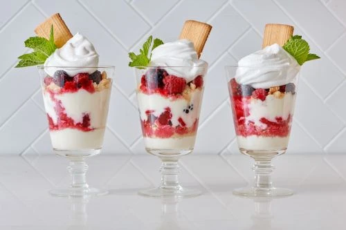 Three glass dessert cups filled with layers of berries, cream, and cookies, topped with mint, against a white brick background.