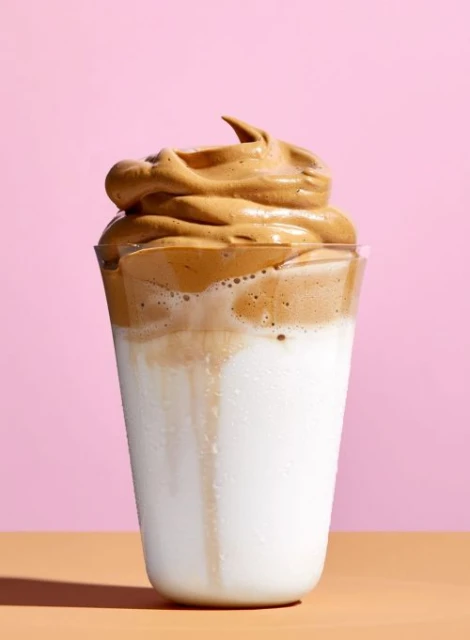 Glass of Dalgona coffee with frothy coffee topping over white milk, set against a pink background on a tan surface. Food and beverage.
