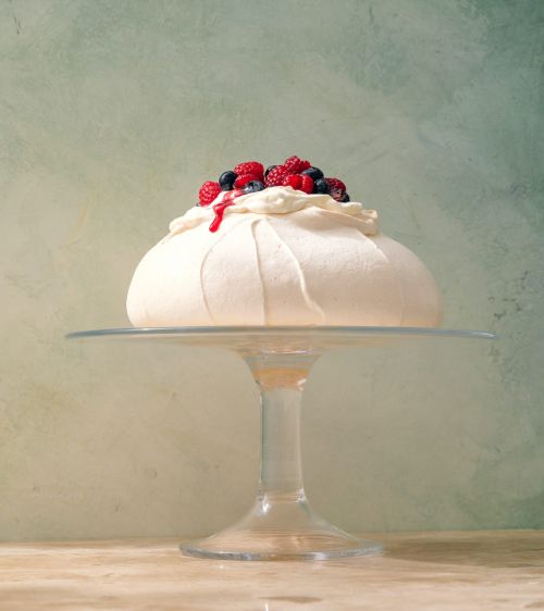 Pavlova cake with fresh raspberries and blueberries on a glass cake stand against a textured green and cream background.