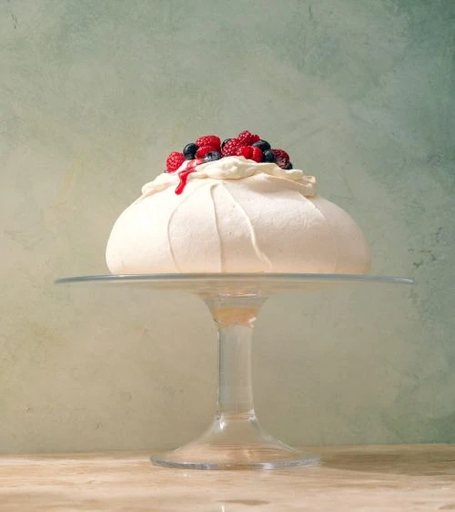 Pavlova cake with fresh raspberries and blueberries on a glass cake stand against a textured green and cream background.