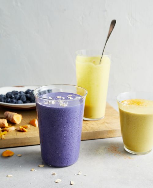 Three smoothie glasses filled with colorful smoothies, blueberries in a bowl, turmeric root, and a spoon on a wooden board.
