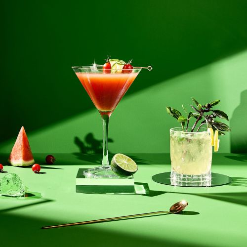 Two cocktails presented with watermelon, lime, basil, and a gold spoon against a graduated green background for a vibrant, refreshing feel.