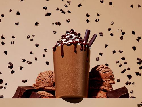 Chocolate milkshake with whipped cream, drizzled chocolate, and chocolate shavings against a beige backdrop with falling chocolate curls.