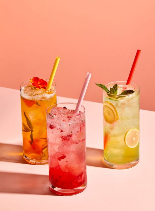 Three colorful cocktails in tall glasses with straws, garnished with lemon, mint, and edible flowers, set against a peach background.