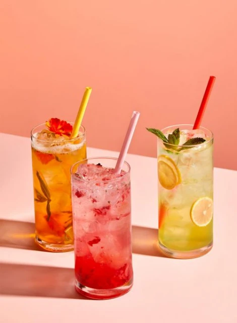 Three colorful cocktails in tall glasses with straws, garnished with lemon, mint, and edible flowers, set against a peach background.