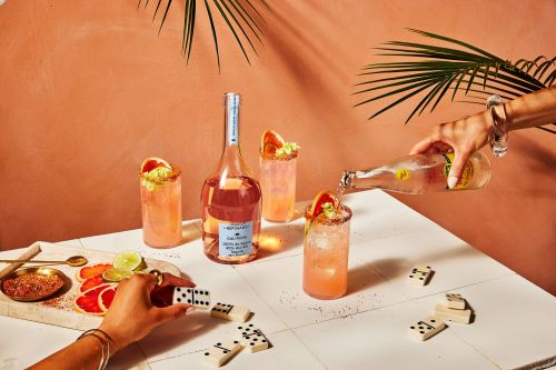 Cocktail glasses with grapefruit slices on a white table, a bottle being poured, dominoes, palm fronds, and an orange background.