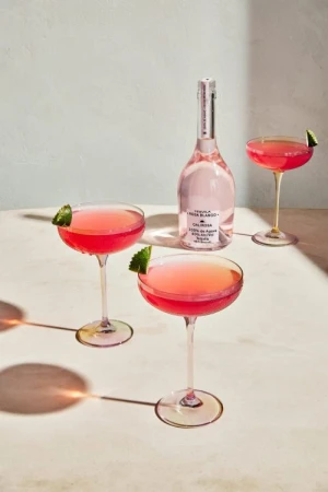 Three pink cocktails in coupe glasses alongside a bottle of Calirosa Tequila on a light surface with shadows, garnished with lime.