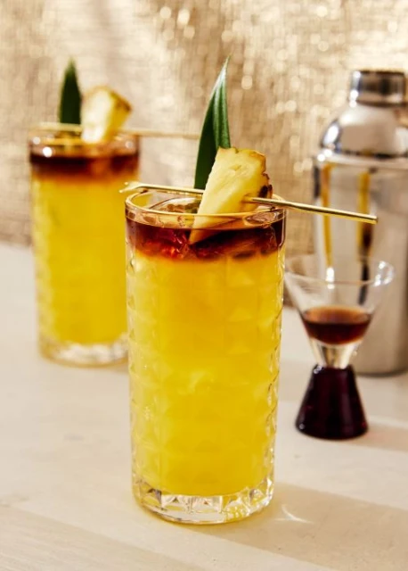 Two pineapple cocktails garnished with pineapple wedges and leaves are in a studio with a gold, shiny background, creating a party mood.