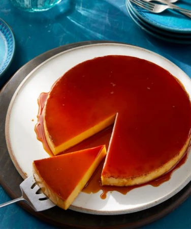 Flan dessert on a white plate, served with a slice removed. The custard dessert is topped with caramel and garnished with plates in the background.