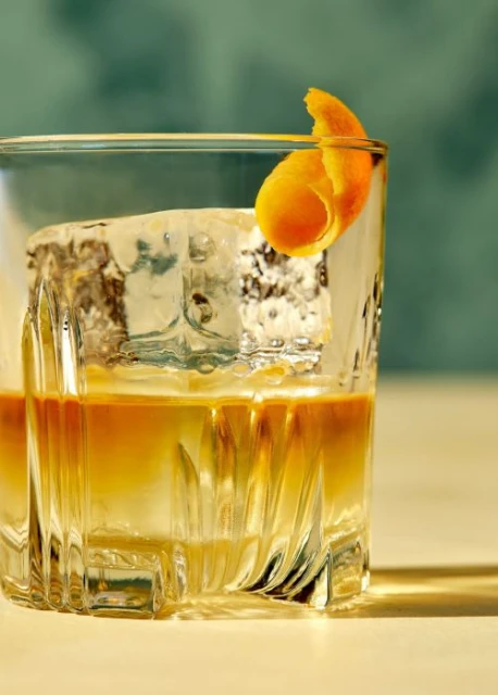 A clear glass with an old-fashioned cocktail made with whiskey, ice, and an orange peel garnish sits on a light surface.
