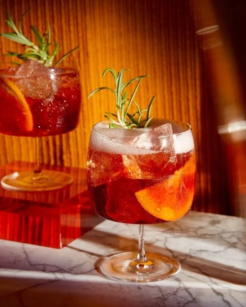 Two stem glasses filled with red cocktails garnished with rosemary and orange slices, arranged on a marble surface in warm lighting.