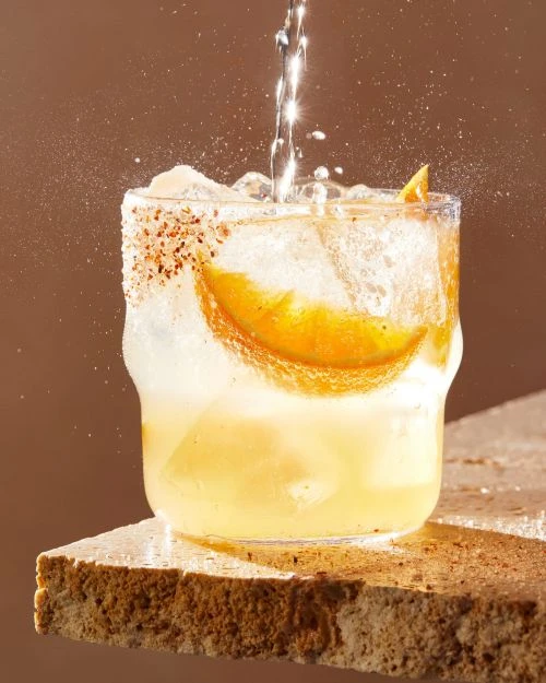 Refreshing mixed drink with orange slice and ice in a glass, being poured with water and garnished, set on a stone surface.