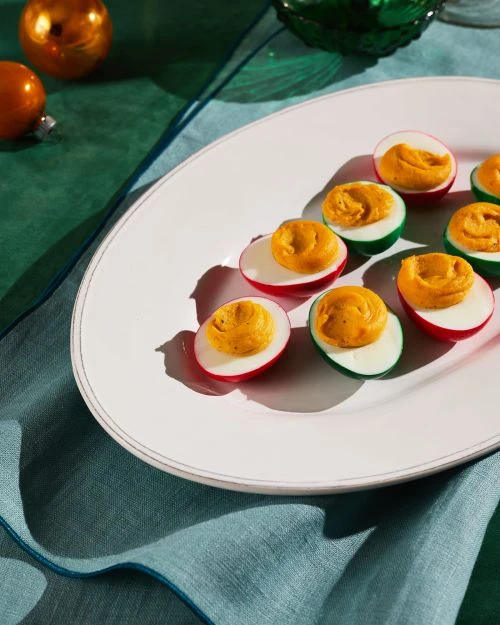 Plate of deviled eggs dyed red and green for a festive appetizer, set on a teal linen cloth with gold ornament in background.