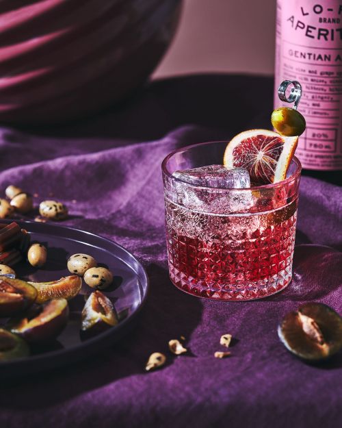 A pink aperitif cocktail with ice and an orange slice, served with plums and nuts on a purple linen surface in a moody setting.