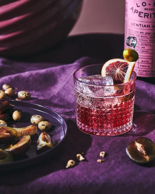 A pink aperitif cocktail with ice and an orange slice, served with plums and nuts on a purple linen surface in a moody setting.