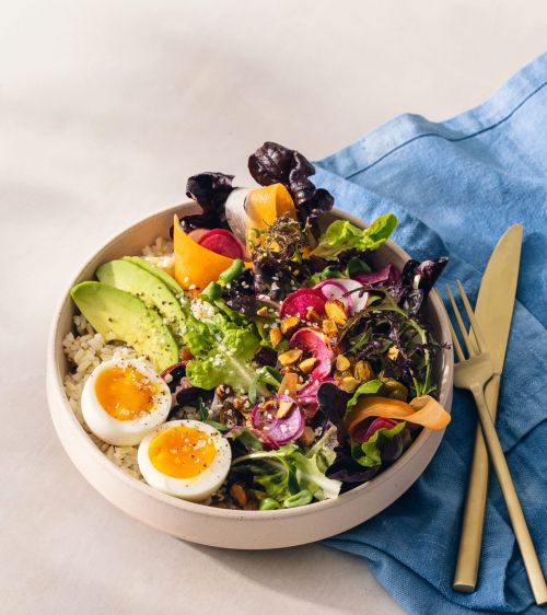 A fresh salad bowl filled with mixed greens, sliced avocado, boiled eggs, and radishes, with golden silverware and a blue cloth.