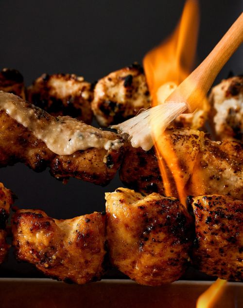 Close-up of grilled chicken skewers, some coated in sauce, with a culinary torch flame brushing over the chicken, on a dark background.