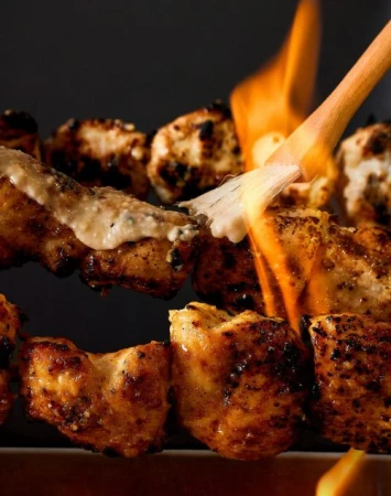 Close-up of grilled chicken skewers, some coated in sauce, with a culinary torch flame brushing over the chicken, on a dark background.