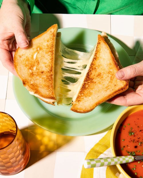Hands holding a grilled cheese sandwich with melted cheese oozing on a green plate, next to tomato soup and a glass of orange juice.