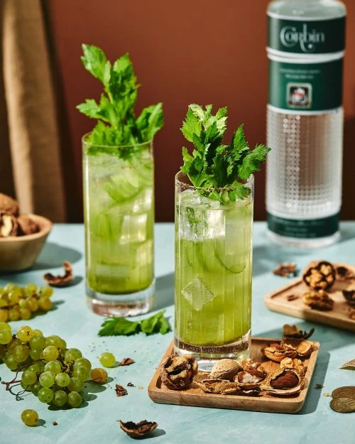 Two green cocktails garnished with celery sit next to walnuts and grapes on a light blue table with a brown background, product shot.