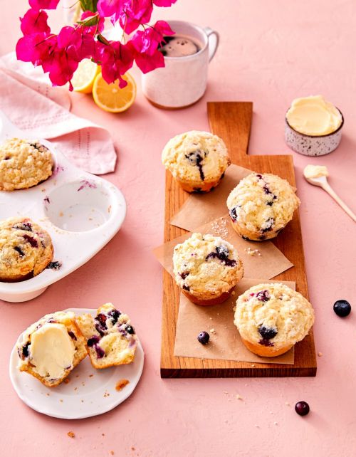 Overhead shot of blueberry muffins displayed on a wooden board and muffin tin next to a cup of coffee, bougainvillea and lemons.