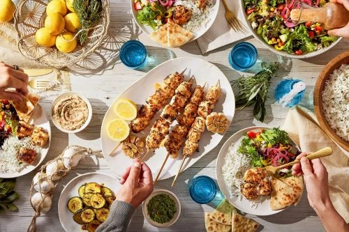 Overhead view of a picnic table laden with grilled chicken skewers, fresh lemons, and sides, surrounded by hands reaching for food.