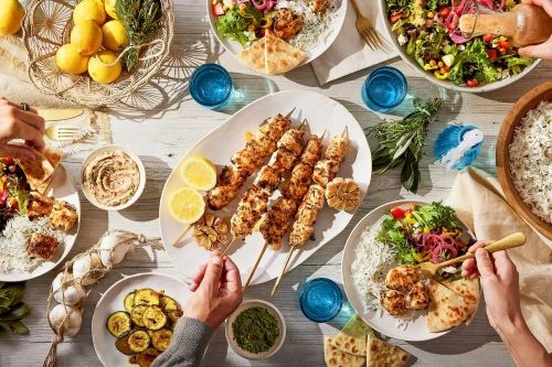 Overhead shot of a dinner party with chicken skewers, salad, rice, bread, lemons, garlic, and other foods arranged on a white table.
