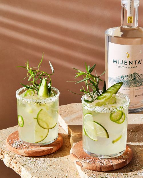 Two margarita cocktails garnished with lime slices and herbs sit on wooden coasters next to a bottle of Mijenta Tequila Blanco.