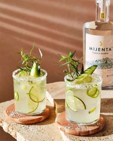 Two margarita cocktails garnished with lime slices and herbs sit on wooden coasters next to a bottle of Mijenta Tequila Blanco.