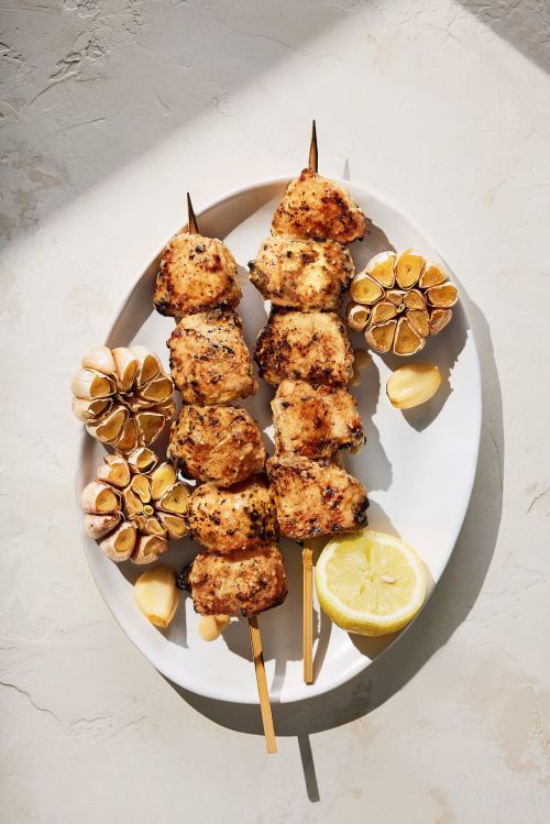 Overhead shot featuring chicken skewers on a white plate, accompanied by roasted garlic bulbs and a lemon half, creating a food still life.