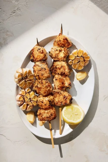 Overhead shot featuring chicken skewers on a white plate, accompanied by roasted garlic bulbs and a lemon half, creating a food still life.