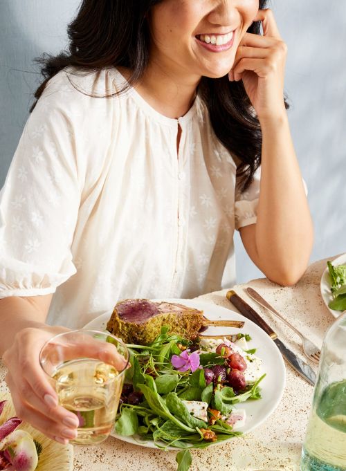 Smiling woman enjoys a healthy meal with lamb chop, salad, and wine at a bright table setting, creating a fresh and inviting scene.