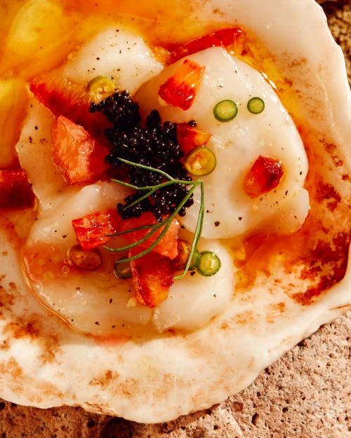 Overhead shot of scallops served with caviar, tomato, and chili garnish in a shell, presented on a textured brown surface.