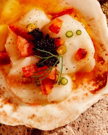 Overhead shot of scallops served with caviar, tomato, and chili garnish in a shell, presented on a textured brown surface.