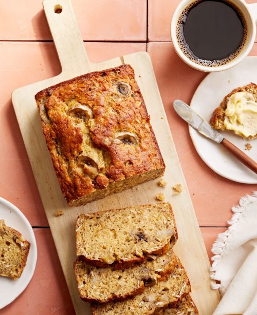 Overhead shot of a sliced banana bread loaf on a wooden cutting board alongside a cup of coffee and buttered slice of bread.