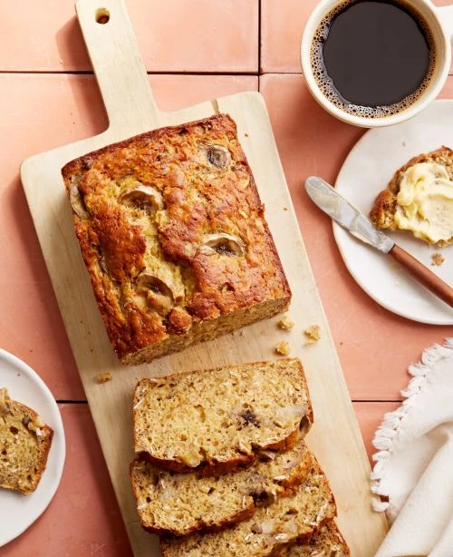 Overhead shot of a sliced banana bread loaf on a wooden cutting board alongside a cup of coffee and buttered slice of bread.