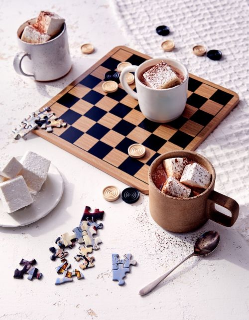 Overhead shot of hot chocolate with marshmallows in mugs, puzzle pieces, and checkerboard on a white textured surface indoors.
