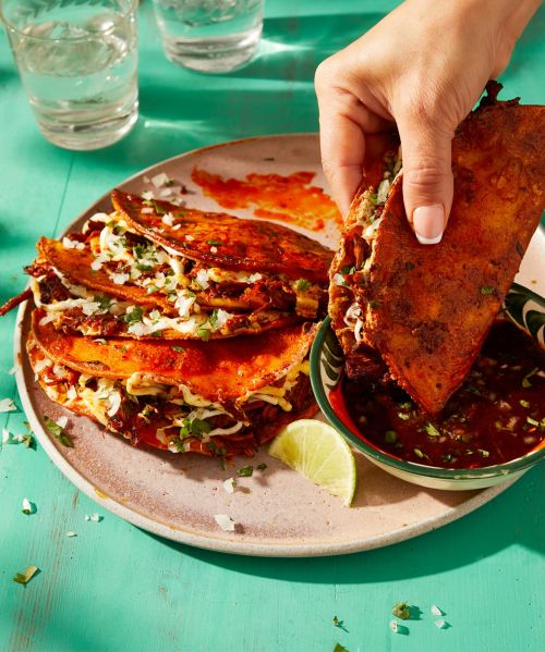 A hand dips a birria taco into a bowl of consommé, with a plate of tacos and glasses of water on a turquoise table, food photography.