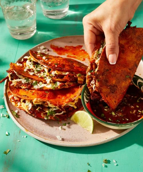 A hand dips a birria taco into a bowl of consommé, with a plate of tacos and glasses of water on a turquoise table, food photography.
