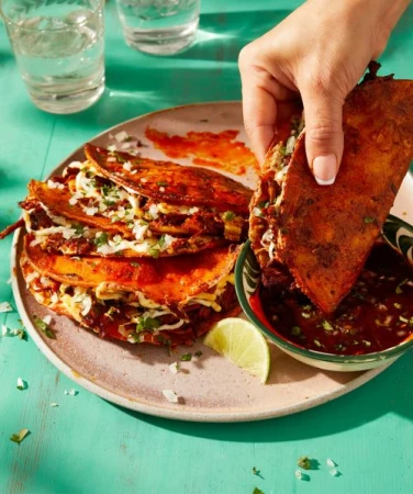 A hand dips a birria taco into a bowl of consommé, with a plate of tacos and glasses of water on a turquoise table, food photography.