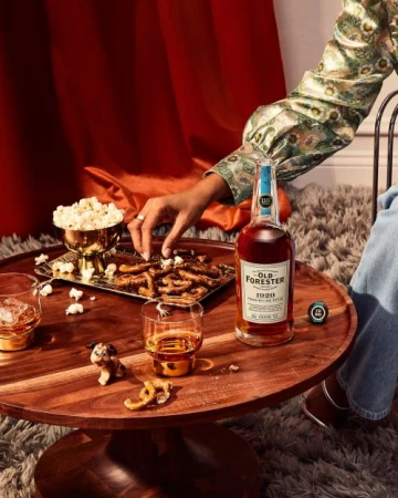 Still life with Old Forester bourbon bottle, drinks, snacks on wood table with a hand reaching for pretzels, against a red curtain.