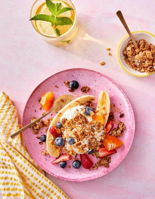 Overhead shot of a banana split with toppings, a glass of iced tea, and a bowl of granola on a pink surface, food photography.