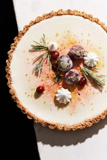 Overhead shot of a tart topped with white cream, cherries, meringues, and rosemary sprigs, sprinkled with gold dust. Food and beverage.