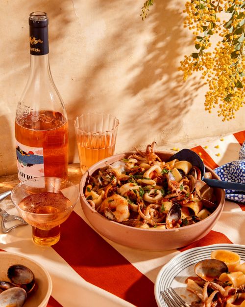 A close-up of a bowl of seafood pasta with a bottle of rose wine and a glass on a striped tablecloth, with floral accents.